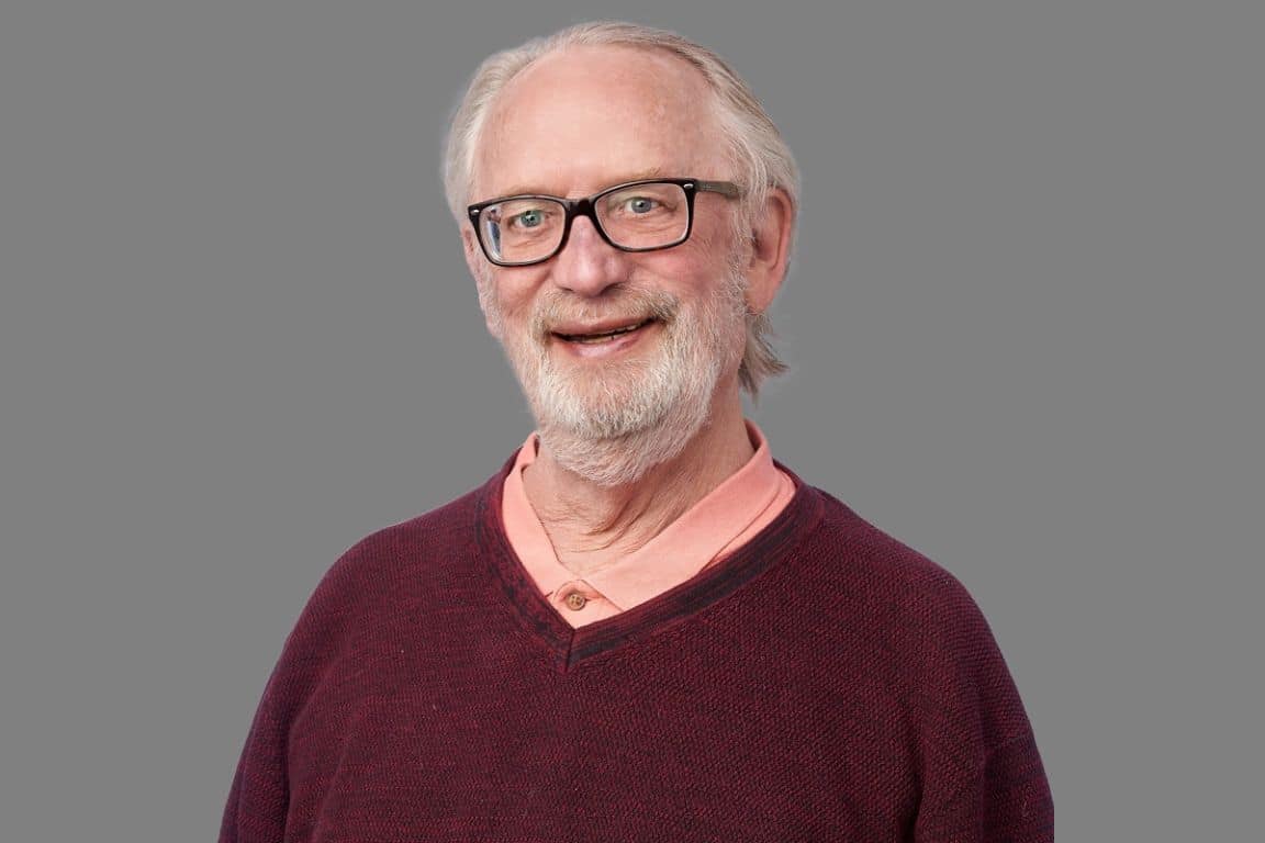 A man wearing a knit sweater, glasses, and a white beard, smiling at the camera.