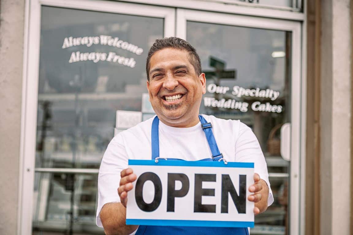 Man smiling and holding an "OPEN" sign in front of a shop with text advertising specialty wedding cakes.