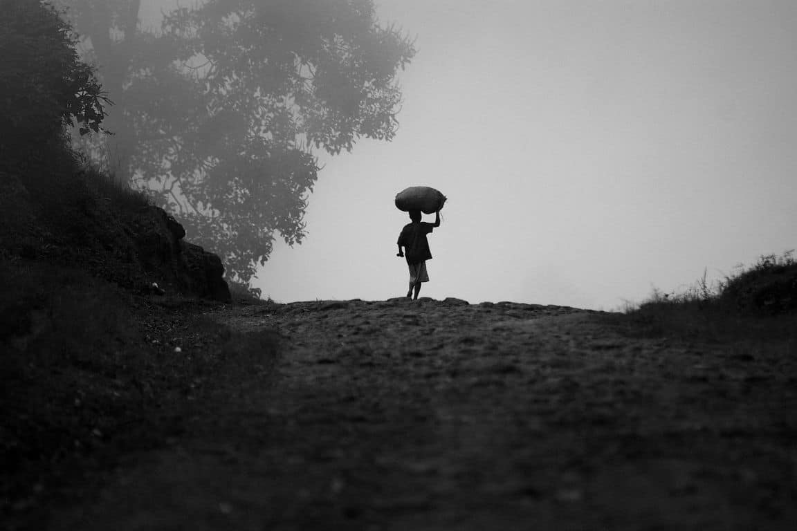 Silhouette of a child walking on a foggy path, carrying a large bag on their head.