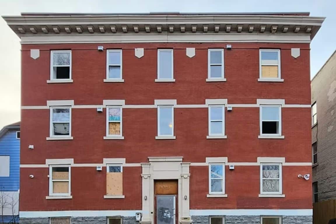 A three-story red brick apartment building with boarded-up windows and doors, symmetrical facade, and stone detailing around the entrance and windows.