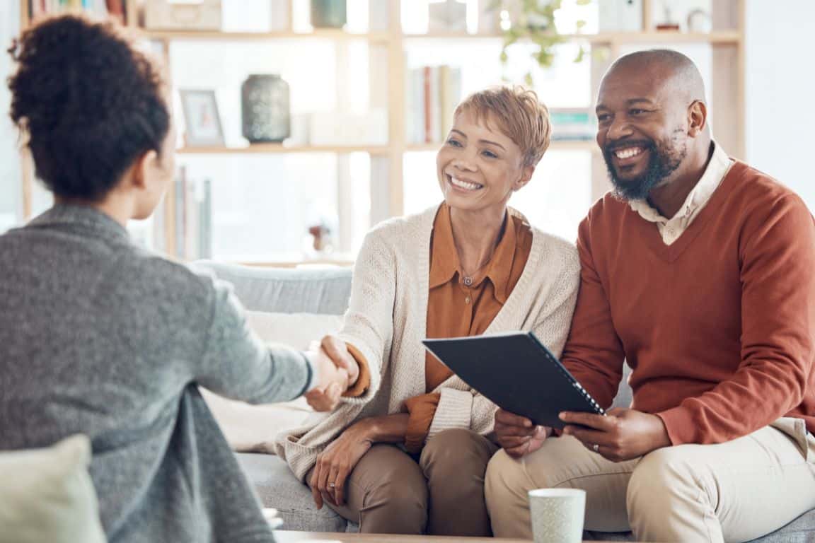 A woman shakes hands with another woman while a man sits beside them holding a folder; all three are seated indoors and appear to be in a business or consultation meeting.