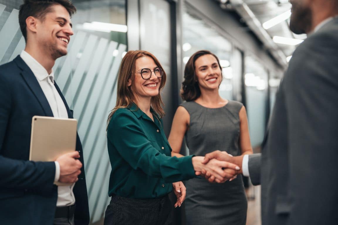 Business team in a modern office: smiling colleagues watch as a woman in green shakes hands with a suited man, conveying warmth and success.
