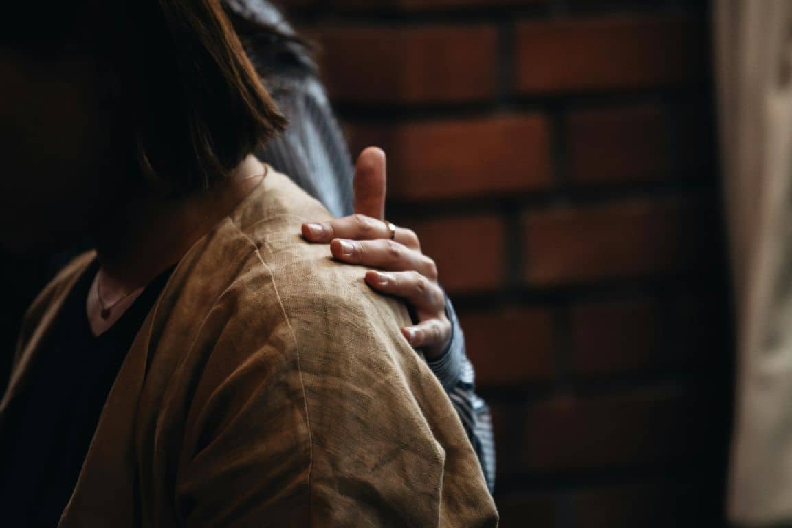 A comforting hand rests on a woman’s shoulder as someone embraces her